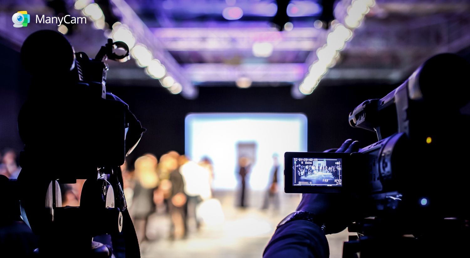 Silhouette of a video camera filming a blurred group of people on stage under bright lights.